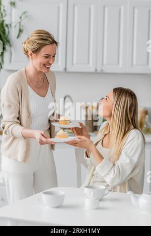 Schließen Sie süße Schwestern in trendigen, pastellfarbenen Cardigans, die Teller mit Cupcakes halten und lächeln, sich verbinden Stockfoto