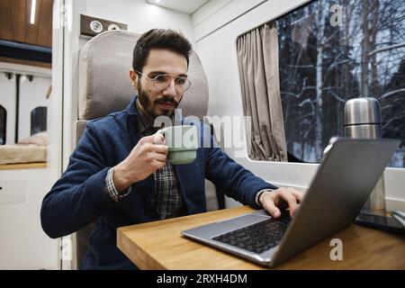 Porträt eines jungen Mannes, der mit Laptop im Wohnwagen arbeitet und Morgenkaffee genießt, Kopierraum Stockfoto