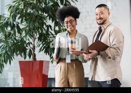 Fröhliche interrassische Büroangestellte, die während der Kaffeepause die Kamera betrachten, Unternehmensvielfalt Stockfoto