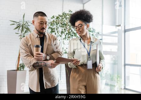 asiatischer Mann zeigt Ordner mit Startup-Plan für afroamerikanische Geschäftsfrau in der Bürohalle Stockfoto