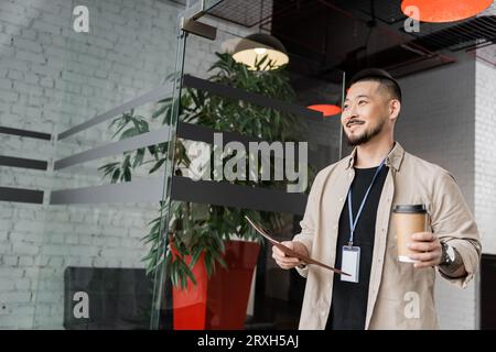 Fröhlicher asiatischer Geschäftsmann mit tätowiertem Lächeln und einem Spaziergang ins Büro mit Ordner und Kaffee zum Mitnehmen Stockfoto