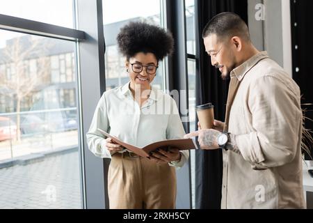 afroamerikanische Frau zeigt Startup-Projekt im Ordner an glücklichen asiatischen Geschäftsmann im Büro Stockfoto