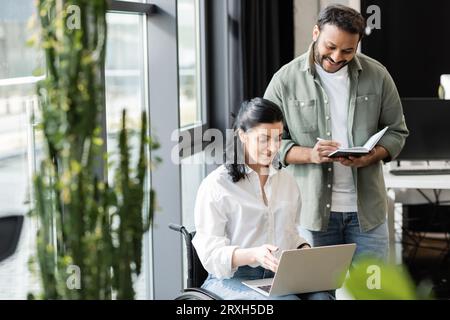 Glücklicher indischer Mann mit Notebook in der Nähe einer behinderten Geschäftsfrau auf Rollstuhl mit Laptop im Büro Stockfoto