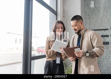 Glückliche Geschäftsfrau zeigt einem asiatischen Kollegen in einem modernen Büro ein Tablet mit Startup-Projekt Stockfoto