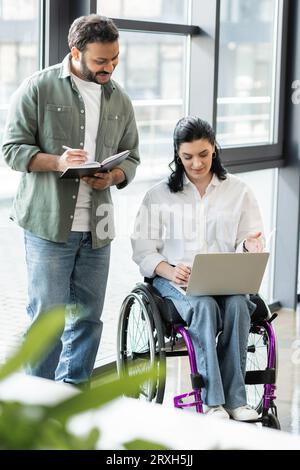 Glücklicher indischer Mann, der Notizen in der Nähe einer behinderten Geschäftsfrau im Rollstuhl macht, die einen Laptop im Büro benutzt Stockfoto