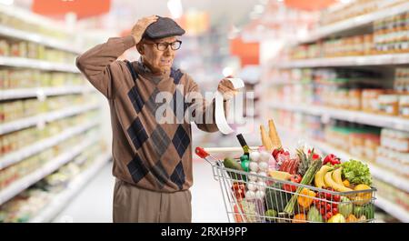 Ein älterer Mann mit einem Einkaufswagen, der sich eine Rechnung anschaut und seinen Kopf in einem Supermarkt ungläubig hält Stockfoto