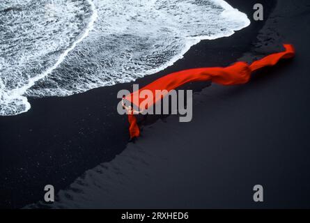 Frau in fließendem Rot an einem schwarzen Sandstrand am Rande der Brandung in Island; Island Stockfoto
