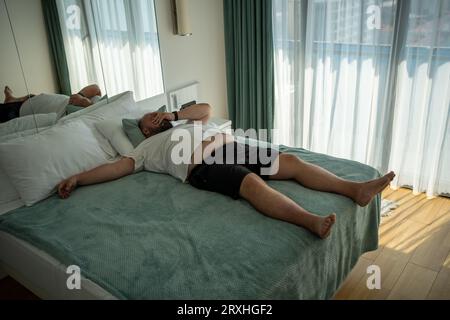 Fette übergroße Männer leiden an Hitze. Fauler, übergewichtiger Mann liegt auf dem Bett und fühlt sich müde im Zimmer Stockfoto