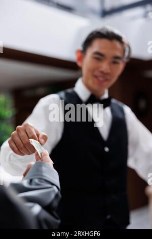 Foto, auf dem ein glücklicher asiatischer Pagenjunge gezeigt wird, der ein Bargeld vom Touristen erhält, nachdem er mit dem Gepäck in der Hotellobby geholfen hat. Gäste, die einchecken, erhalten freundlichen Service und geben dem Concierge Geld. Stockfoto