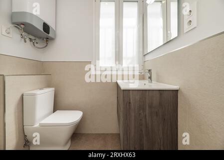 Badezimmer mit einem neuen Holzschrank mit weißem Porzellanwaschbecken und rahmenlosem quadratischen Spiegel, der an der Wand hängt, Warmwasserbereiter auf der Toilette und weiß Stockfoto