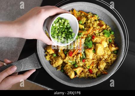 Frau fügt geschnittene grüne Zwiebeln zu Reis mit Fleisch und Gemüse in der Pfanne hinzu, Draufsicht Stockfoto