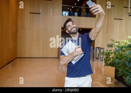 Lächelnder Büroangestellter, der während der Pausen in der Bürohalle per Videoanruf mit einem Freund spricht Stockfoto