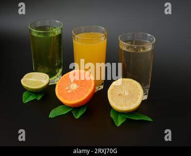 Drei große Gläser mit verschiedenen Zitrussäften auf schwarzem Hintergrund in einer Reihe, neben den Hälften von Limette, Orange und Zitrone mit Blättern. Nahaufnahme. Stockfoto