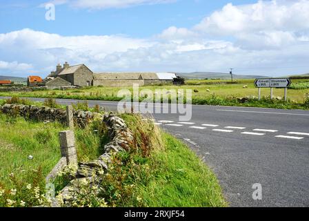 Ländliche Kreuzung auf halbem Weg zwischen Kirkwall und Stromness auf Orkney Island, Schottland. Auf der anderen Straßenseite befinden sich eine historische Steinscheune und andere Gebäude. Stockfoto