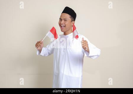 Porträt eines attraktiven asiatischen muslimischen Mannes in einem weißen Hemd mit einer Kopfbedeckung, die die indonesische Flagge hält, während sie seine Faust hebt und den Erfolg feiert. Isolierte Bildgebung Stockfoto