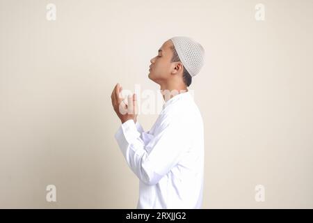 Porträt eines attraktiven asiatischen muslimischen Mannes in weißem Hemd mit Kopfbedeckung, der ernsthaft mit erhobenen Händen betet. Isoliertes Bild auf grauem Hintergrund Stockfoto