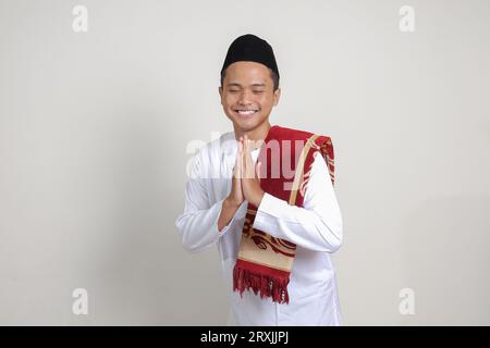 Porträt eines attraktiven asiatischen muslimischen Mannes in weißem Hemd mit Skullcap mit entschuldigender und willkommener Handgeste. Isoliertes Bild auf grauem Hintergrund Stockfoto