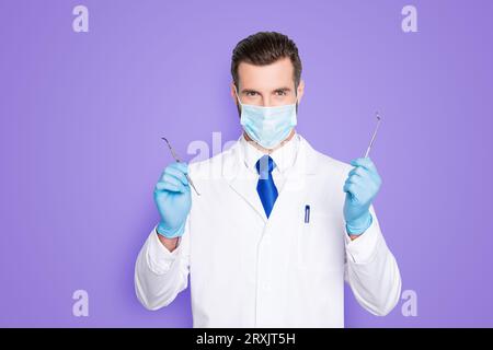 Porträt eines erfolgreichen gutaussehenden Zahnarztes in weißem Laborkittel mit blauer Krawatte, die Werkzeuge für die Zahnbehandlung mit den Händen hat, anschauen Stockfoto