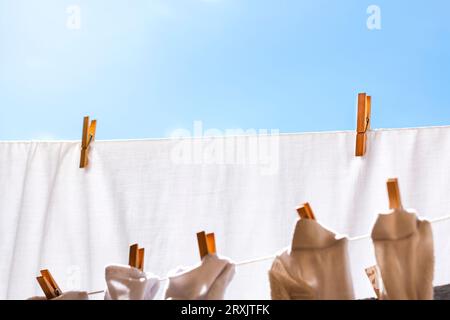 Von unten hängen weiße und graue Kleidungsstücke an Wäscheleinen, während sie im Sommer in Sonnenlicht trocknen und mit Holzwäschespins daran befestigt werden Stockfoto