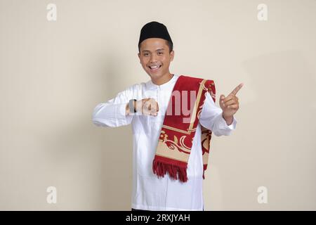 Porträt eines attraktiven asiatischen muslimischen Mannes in weißem Hemd, der auf eine Armbanduhr schaut, die mit dem Finger nach vorne zeigt. Isoliertes Bild auf grauem Hintergrund Stockfoto