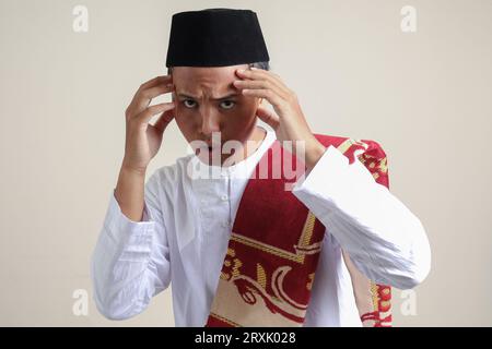 Porträt eines attraktiven asiatischen muslimischen Mannes in weißem Hemd mit Kopfbedeckung, die sich gutaussehend und selbstbewusst anfühlt. Seine Schläfen berühren, um einen Stil zu kreieren. Isoliert im Stockfoto