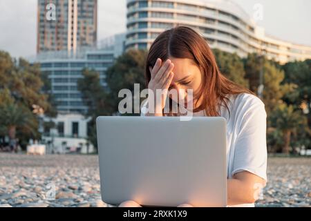 Müde Frau Freiberuflerin im Stress, die draußen am Laptop arbeitet und ihren Kopf mit der Hand hält. Frustrierte und erschöpfte digitale Nomadin mit Kopfschmerzen Stockfoto