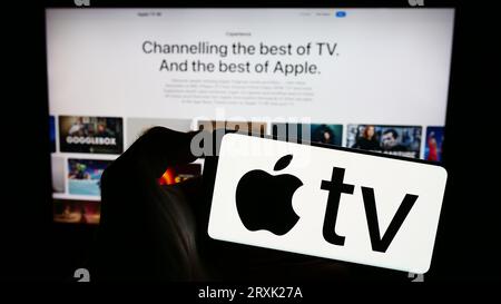 Person, die ein Mobiltelefon mit dem Logo des Video-Streaming-Dienstes Apple TV plus auf dem Bildschirm vor der Business-Webseite hält. Konzentrieren Sie sich auf das Display des Telefons. Stockfoto