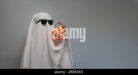 Lustiges Halloween-Kinderkonzept, kleines süßes Kind mit weiß gekleidetem Kostüm halloween-Geister, der orangefarbenen Kürbisgeister zur Hand hält Stockfoto