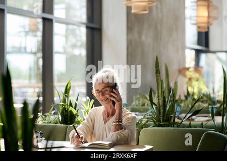Ältere Frau, die sich während ihrer Arbeit im Café mit dem Handy unterhalten und Notizen in ihrem Notizblock macht Stockfoto