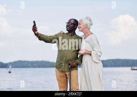 Ein multiethnisches Seniorenpaar macht Selfie auf dem Smartphone gegen den See Stockfoto