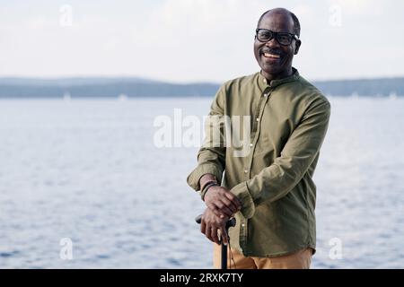 Porträt eines afroamerikanischen glücklichen älteren Mannes, der vor der Kamera lächelt, während er in der Nähe des Sees spaziert Stockfoto