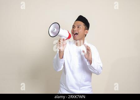 Porträt eines attraktiven asiatischen muslimischen Mannes in weißem Hemd mit Skullcap-sprechendem lauter mit Megaphon, das Produkt fördert. Werbekonzept. Isoliert Stockfoto