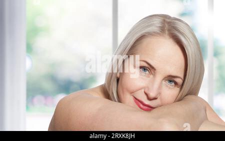 Glückliche schöne blonde Frau mittleren Alters zeigt ihr perfekt gepflegtes Gesicht und sitzt entspannt am Fenster. Realistische Bilder. Stockfoto