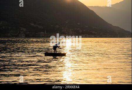 Bei Sonnenuntergang, inmitten der Berge, fischt ein Mann in einem kleinen Boot in der Bucht. Sonnenreflexion. Querformat. Stockfoto