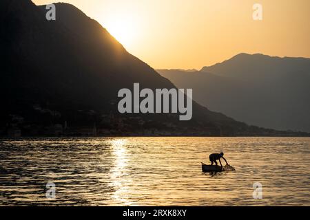 Bei Sonnenuntergang fischt ein Mann in einem kleinen Boot in der Bucht. Silhouetten in der goldenen Stunde. Dramatische Szene. Berge im Hintergrund. Querformat. Stockfoto