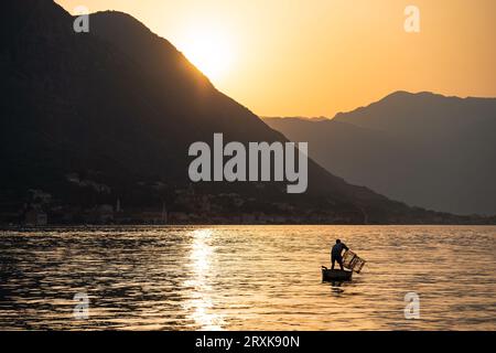 Bei Sonnenuntergang, inmitten der Berge, fischt ein Mann in einem kleinen Boot in der Bucht. Silhouetten in der goldenen Stunde. Dramatische Szene. Stockfoto