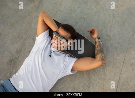 Stilvoller tätowierter Mann, der sich auf dem Skateboard entspannt und Musik auf den Ohrpolstern hört, sich im Skatepark entspannt, Blick von oben Stockfoto