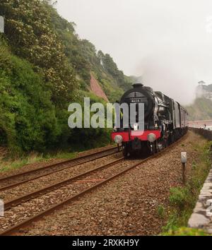 An einem bewölkten Tag fährt die LMS Royal Scot der Klasse 46115 Scots Guardsman entlang der Seemauer in Teignmouth mit der Royal Herzogtum Bahnfahrt. Stockfoto