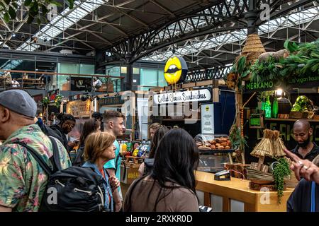 Leute, die sich anstellen, um Lebensmittel in den Lebensmittelgeschäften des Old Spitalfields Market, London, E1, zu kaufen Stockfoto