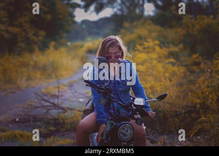 Schöne Frau mit blauer Jeansjacke sitzt auf Enduro-Motorrad vor farbenfrohem natürlichem Hintergrund Stockfoto