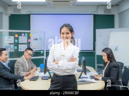 Porträt lächelnde moderne Geschäftsfrau, die im Büro bei einem Teammeeting steht. Selbstbewusste junge asiatische Geschäftsfrau mit Kollegen im Sitzungssaal. Treffen Stockfoto