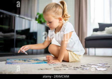 Das kleine blonde Mädchen sitzt zu Hause auf dem Teppich und sammelt Rätsel Stockfoto