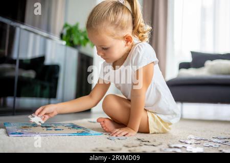 Das kleine blonde Mädchen sitzt zu Hause auf dem Teppich und sammelt Rätsel Stockfoto