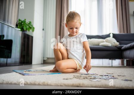 Das kleine blonde Mädchen sitzt zu Hause auf dem Teppich und sammelt Rätsel Stockfoto