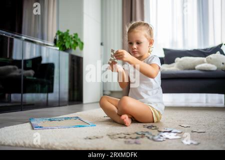 Das kleine blonde Mädchen sitzt zu Hause auf dem Teppich und sammelt Rätsel Stockfoto