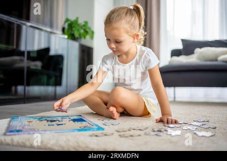 Das kleine blonde Mädchen sitzt zu Hause auf dem Teppich und sammelt Rätsel Stockfoto