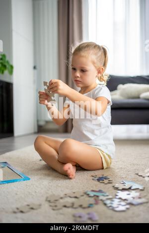 Das kleine blonde Mädchen sitzt zu Hause auf dem Teppich und sammelt Rätsel Stockfoto