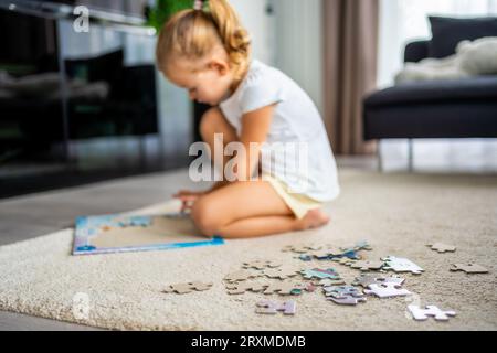 Das kleine blonde Mädchen sitzt zu Hause auf dem Teppich und sammelt Rätsel Stockfoto