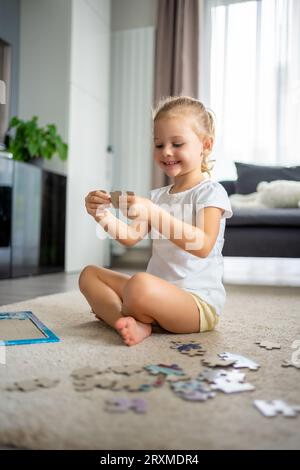 Das kleine blonde Mädchen sitzt zu Hause auf dem Teppich und sammelt Rätsel Stockfoto