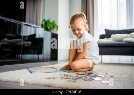 Das kleine blonde Mädchen sitzt zu Hause auf dem Teppich und sammelt Rätsel Stockfoto